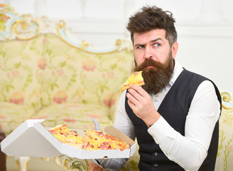 Man with beard and mustache holds delivered box with tasty fresh hot pizza. Macho in classic clothes hungry, on strict face, holds slice of pizza, eats, interior background. Pizza delivery concept.