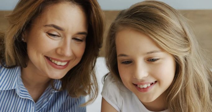 Close up of the pretty young mother and her cute teen daughter lying in the bedroom in the morning and watching something on a laptop computer. Weekend. Inside