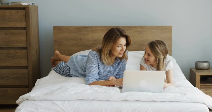 Cheerful smiled mother and litle daughter resting after waking up in the morning on the unmade bed in front of the laptop screen. Indoors