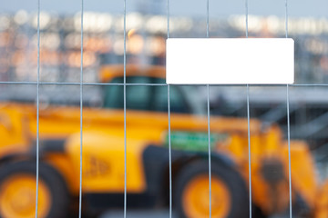 White empty mockup plate with copyspace fixed on a metal grid. Behind the fence in the background is a yellow construction truck. Empty blank sign on blurry background. Advertising concept or logotype