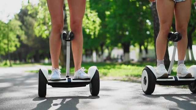 Two girls in short shorts ride the camera and smile at the self-balancing scooter in the Park