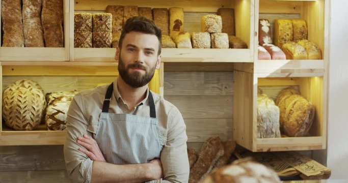 Portrait of the young Caucasian male bread seller in the apron looking at the side and then smiling to the camera in the bakery shop. Indoor