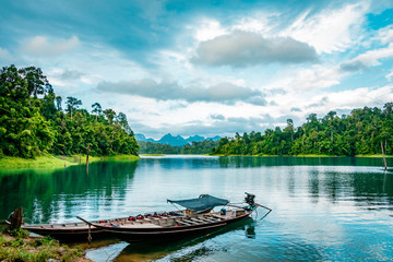 Naklejka na ściany i meble Wooden Thai traditional long-tail boat on a lake with mountains and rain forest in the background during a sunny day at Ratchaprapha Dam at Khao Sok National Park, Surat Thani Province, Thailand