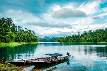 Obraz premium Wooden Thai traditional long-tail boat on a lake with mountains and rain forest in the background during a sunny day at Ratchaprapha Dam at Khao Sok National Park, Surat Thani Province, Thailand
