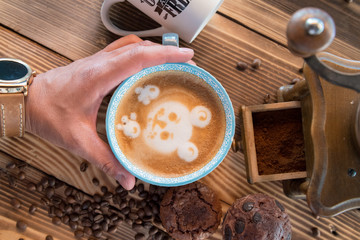 Man hands holding a cup of coffee with foam next to the coffee grinder on wooden table, top view