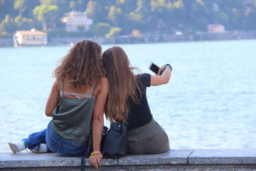 due amiche in riva al lago si fanno un selfie, persone guardano smartphone