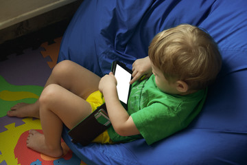 Little boy lies on a bag chair. The child is lying at home on the armchair and looks at the screen of the smartphone. His finger touches the screen and presses on the glass