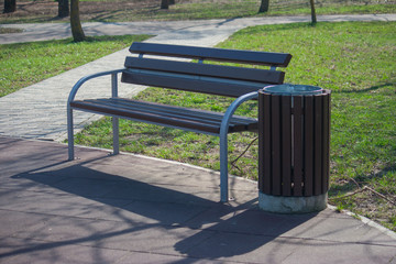 Empty bench in the recreation park. Standard design of a bench for rest and a garbage can near it. Brown wooden seat.