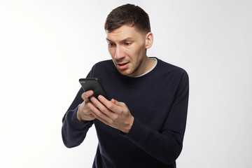 Closeup portrait anxious or shocked young freelancer man looking at phone seeing bad news or photos with disgusting emotion on face isolated over white background. Human emotion, reaction, expression.