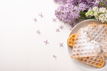 Homemade waffles heart sprinkled with powdered sugar on plate on a white table with flowers. Sweet pastry. Top view