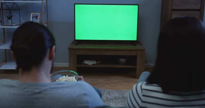 View From The Back On The Couple Watching TV With Green Screen And Eating Popcorn Late In The Evening In The Living Room. Indoor