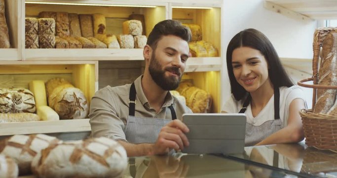 Attractive male and female bread vendors scrolling and watching something on the tablet device while standing at the counter in the bakery shop. Indoor