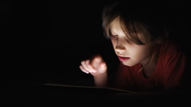 Caucasian girl lying in bed playing tablet in social internet in dark light under blacket. Childhood, communication and technology concept