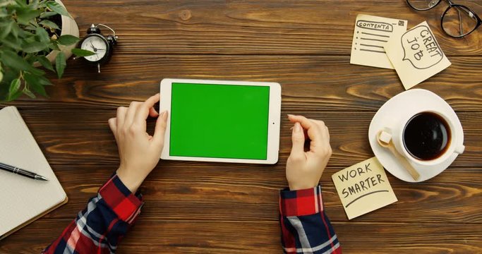 Top view of the white tablet device lying on the wooden desk and woman's Caucasian hands with plaid sleeves scrolling, taping and zooming on its vertical screen near with cup of coffee and notes