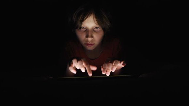 Dolly shot caucasian girl lying in bed playing tablet in social internet in dark light under blacket. Childhood, communication and technology concept
