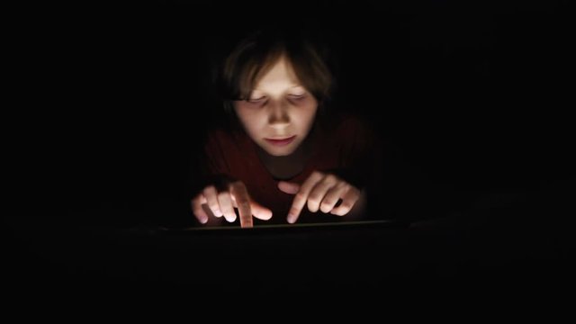 Dolly shot caucasian girl lying in bed playing tablet in social internet in dark light under blacket. Childhood, communication and technology concept