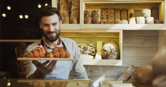 Portrait of the handsome young male baker smiling while posing with croissants on the tray in the shop. Indoors