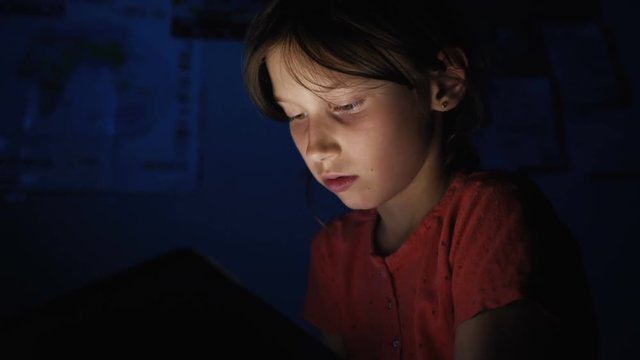 Dolly shot caucasian girl lying in bed playing tablet in social internet in dark blue light under blacket. Childhood, communication and technology concept