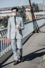 asian businessman in protective mask walking on bridge with coffee to go and looking away, air pollution concept