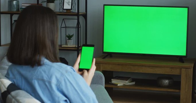 Rear View On The Brunette Womansitting On The Couch In The Living Room, Watching TV With Green Screen And Also Something On The Green Screen Of The Black Smartphone. TV In Focus. Chroma Key. Indoor