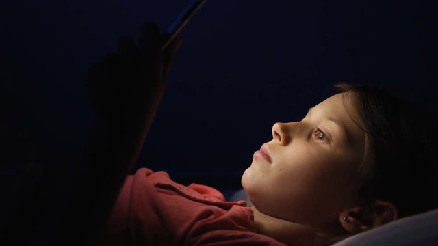 Dolly shot caucasian girl lying in bed playing tablet in social internet in dark blue light under blacket. Childhood, communication and technology concept