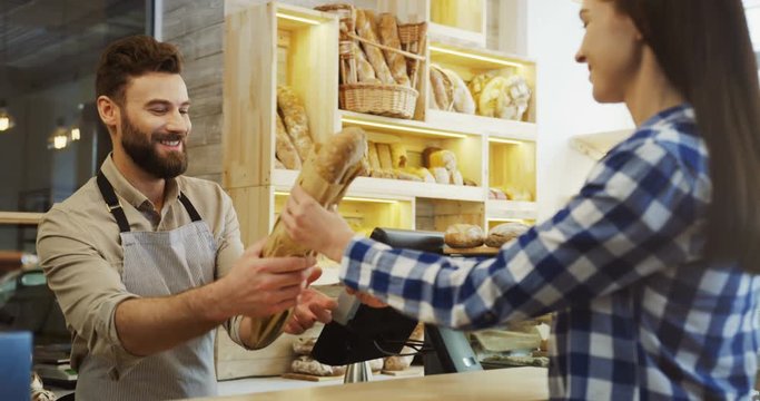 Handsome man, bakery vendor selling baguettes to the young woman and she paying with a credit card. Indoors