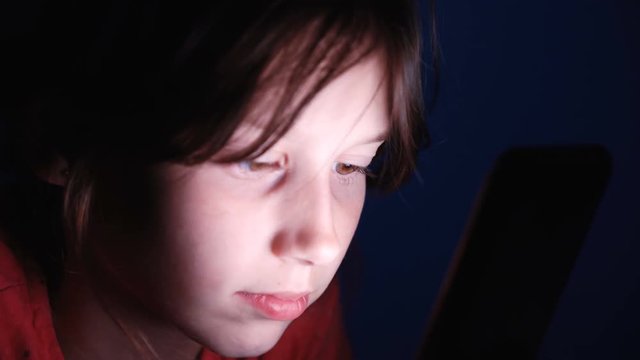 Dolly shot caucasian girl lying in bed playing tablet in social internet in dark blue light under blacket. Childhood, communication and technology concept