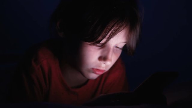 Dolly shot caucasian girl lying in bed playing tablet in social internet in dark blue light under blacket. Childhood, communication and technology concept