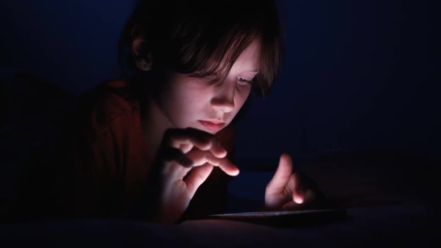Dolly shot caucasian girl lying in bed playing tablet in social internet in dark blue light under blacket. Childhood, communication and technology concept