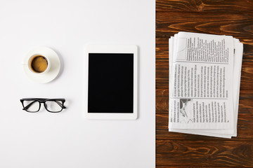 top view of eyeglasses, coffee cup, and digital tablet with blank screen on white and pile of newspapers on wooden background