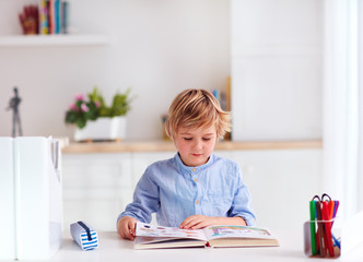 young kid, boy reading a book while sitting at the desk at home