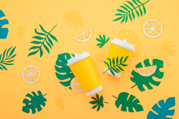 Fototapeta na wymiar Disposable paper cup on a yellow background with tropical leaves and orange slices on a bright yellow background. Colorful summer drink concept with paper cut outs.
