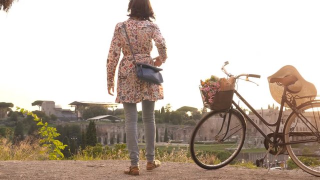 Young woman tourist with bycicle takes pictures of the colosseum in rome at sunset with smartphone. Beautiful stylish dress with large hat, flowers and bread in basket.