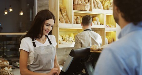 Young beautiful woman standing at the counter in the bakery shop and selling bread to the man, her male co-worker doing something behind her. Indoors - Powered by Adobe