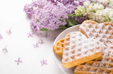 Homemade waffles heart sprinkled with powdered sugar on plate on a white table with flowers. Sweet pastry, breakfast.