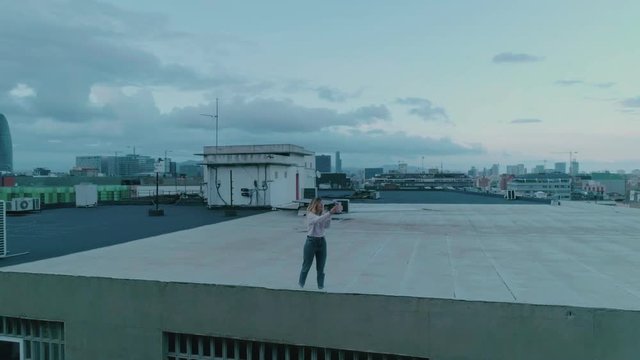 Aerial drone shot of young woman, student or blogger model stand on urban creative rooftop or terrace, makes selfie photos on smartphone or has video chat or call with friends, at beautiful sunset