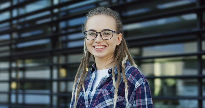 Close up of the pretty Caucasian young woman with dreadlocks and in glasses smiling sincerely to the camera. Outside
