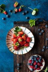 Strawberries, blueberries and raspberries in a cup and scattered on a rustic blue wooden base. Top view