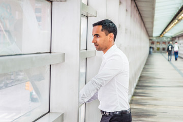 Naklejka na ściany i meble Young Handsome Hispanic American Businessman working in New York, wearing white shirt, black pants, standing on walkway by glass wall inside office building, looking outside, thinking, lost in thought