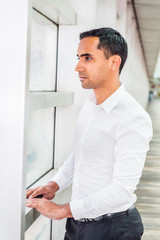 Fototapeta premium Portrait of Young Handsome Hispanic American Man in New York, wearing white shirt, standing by glass wall, looking outside, thinking, lost in thought. Side View..