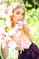Fototapeta premium Charming blond girl enjoying scent of flowering trees. Pretty lady relaxing and breathing fresh aroma of blooming magnolia. Attractive curly-haired young woman walking through the spring garden