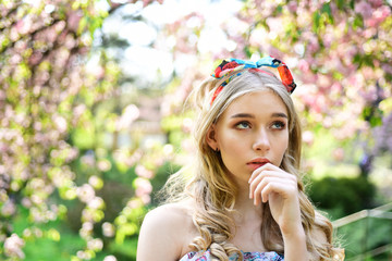 Thoughtful girl in retro outfit with colorful headband dreaming in floral garden, unity with nature concept. Lovely young lady strolling between blooming trees on sunny day