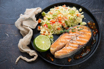 Grilled salmon fillet and cuscus with vegetables on a metal tray, studio shot on a brown stone background
