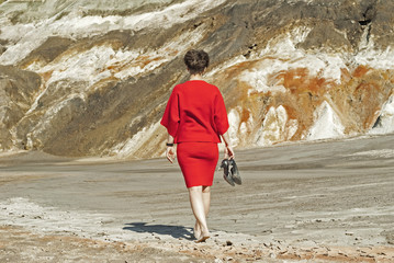 woman in a business red dress, taking off her shoes, walking barefoot into a deserted canyon with her back to the viewer..