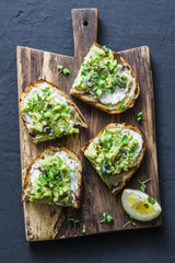 Avocado, cream cheese sandwich with micro greens on a rustic cutting board, top view. Good fats healthy eating concept. Delicious breakfast or snack. On dark background