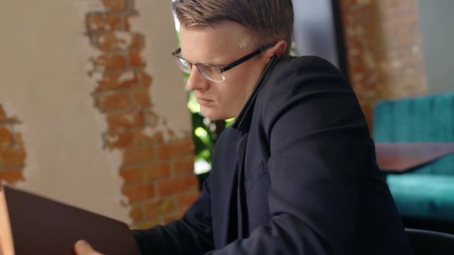 Portrait of young businessman taking notebok and documents from the table and going out in cafe while talking on phone. Concept: business people, documents on desk, cafe interior, phone call