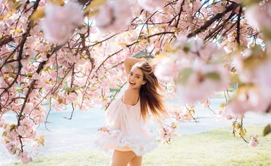 Poster Kersenbloesem Woman with gorgeous smile having fun in blooming garden. Female with slim sexy body on natural background, wellness and fitness concept. Sensual blond girl in short chiffon dress walking in park  © Volodymyr