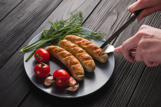 Three grilled sausages on a gray plate on a wooden black background lie also a cherry tomato, greens, garlic, knife, fork. View from above. Place for text and logo.