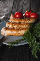 Three grilled sausages on a gray plate on a wooden black background lie also a cherry tomato, greens, garlic, knife, fork. View from above. Place for text and logo.