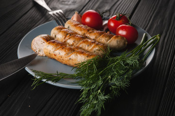 Three grilled sausages on a gray plate on a wooden black background lie also a cherry tomato, greens, garlic, knife, fork. View from above. Place for text and logo.